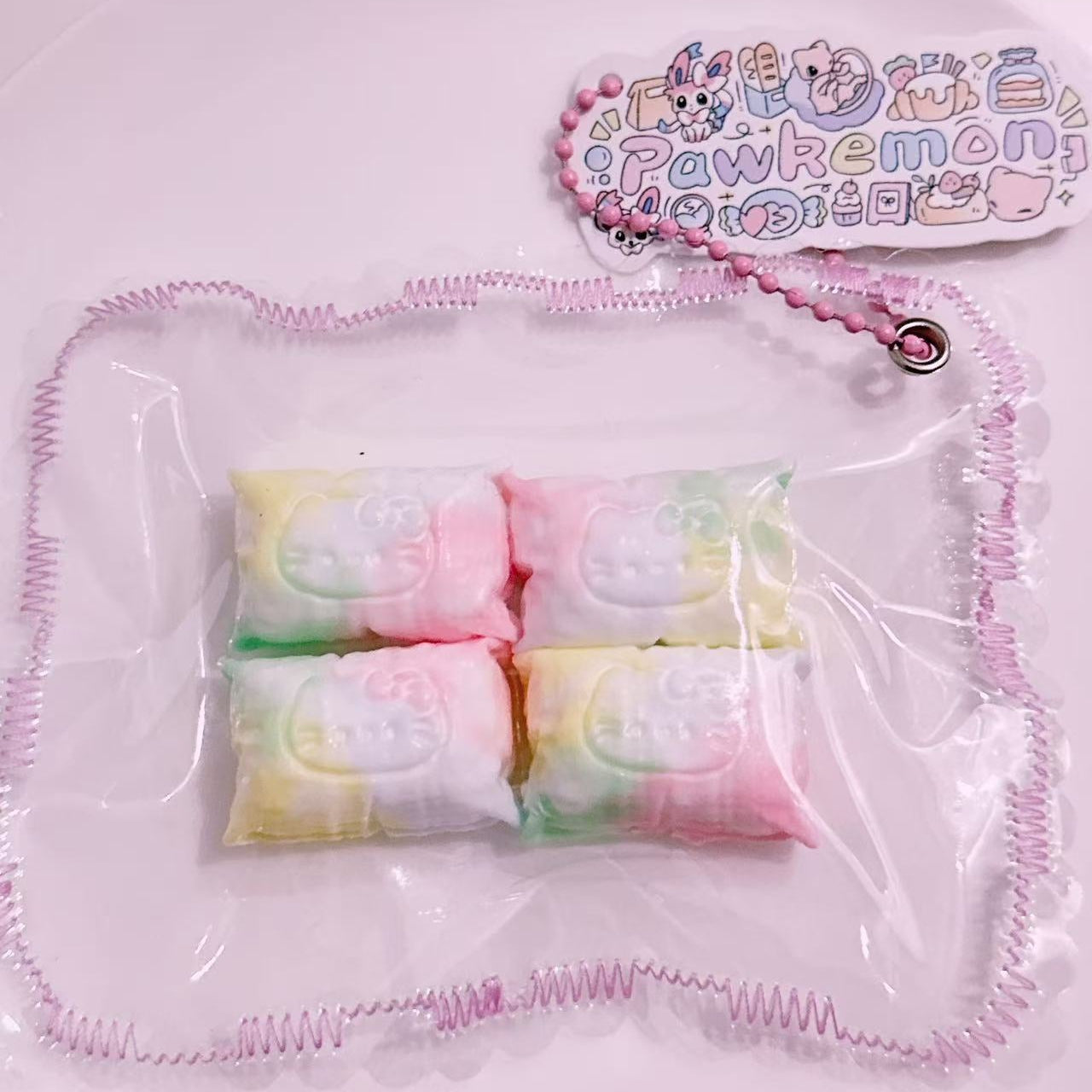 Pastel kitty pillow taba squishy 4-piece set displayed in clear packaging with pink chain tag from Pawkemon. Handmade silicone mini squishy toy with kawaii soft texture.