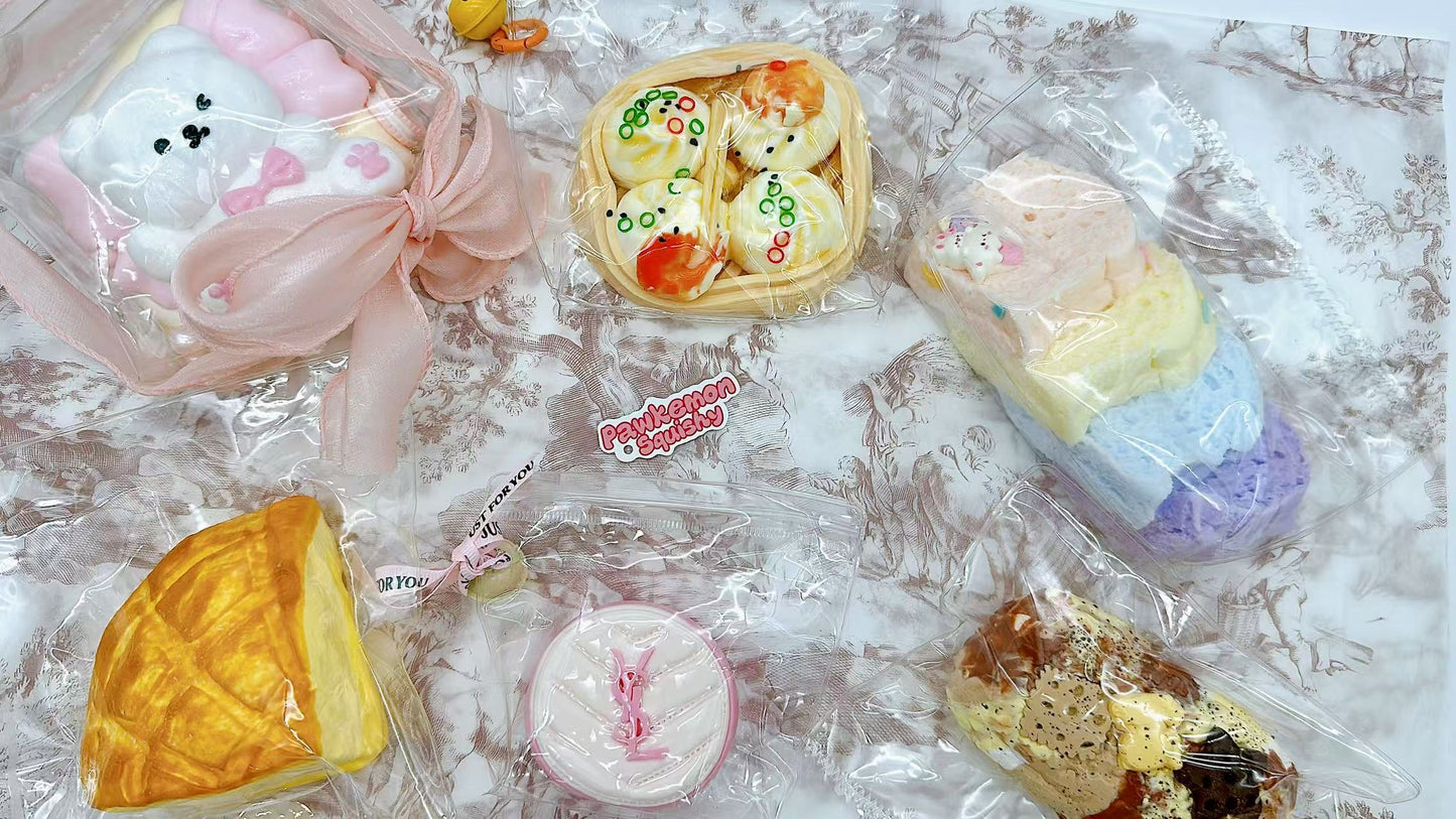 A collection of individually packaged handmade squishies including toast, dim sum, rainbow loaf, and a pink pudding, displayed on a patterned fabric with a soft pink ribbon and Pawkemon Squishy branding.