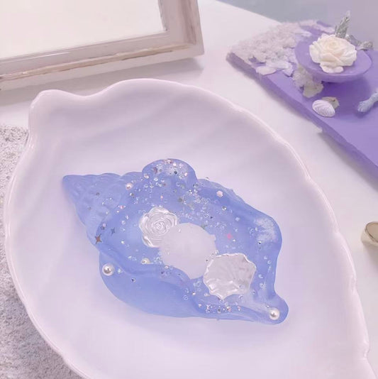 A dreamy Ocean Pearl Conch Taba Squishy in translucent blue, seashell-shaped with sparkling glitter, pearls, and rose details, displayed on a white leaf-shaped plate with soft floral background.
