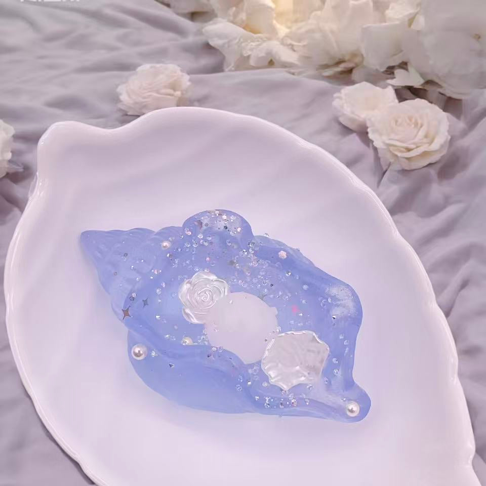Celestial Shell Taba Squishy in dreamy translucent blue, seashell-shaped with pearl, rose, and glitter details, placed on a white shell plate in a soft pastel setting."