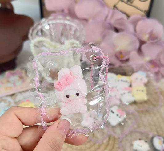 A pink Melody-inspired bunny squishy with a bow, displayed inside a transparent pouch with decorative stitched edges.
