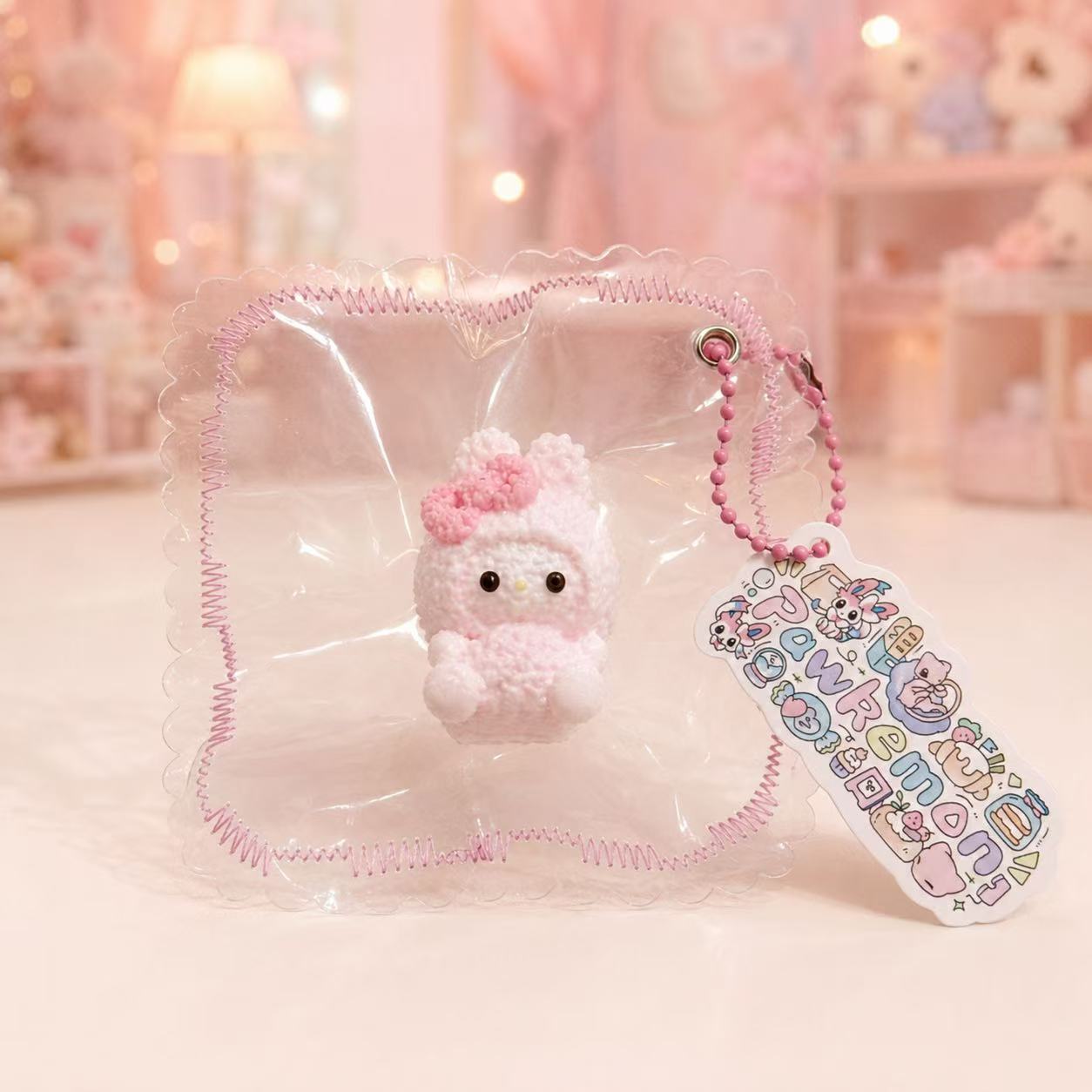 A small pink bunny character squishy inside a clear plastic bag with a pink scalloped edge, shown on a soft pink surface with a blurred boutique background. 