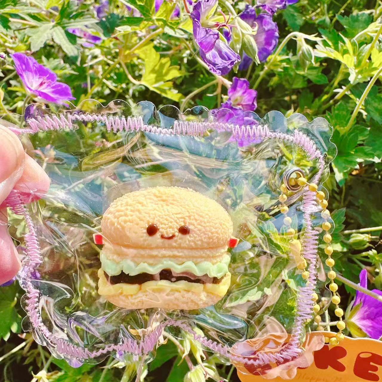 Mini Jellycat Burger Taba Squishy toy in clear PVC bag, cute kawaii burger squishy collectible photographed outdoors with purple flowers background.