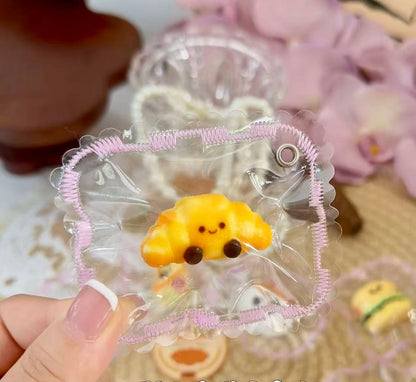 A cute croissant-shaped squishy toy with a smiling face, bright golden color, packaged in a transparent pouch with machine-stitched decorative edges.