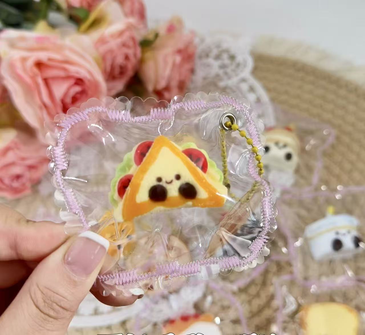 A cute Jellycat Sandwich Taba Squishy with smiling face, layered with cheese, lettuce, and tomato, packaged in a clear pouch with machine-stitched decorative lace edges.