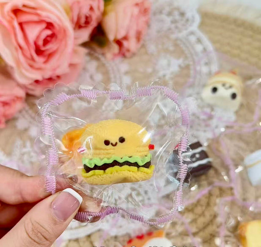 A mini burger-shaped squishy with a smiling face, lettuce, and cheese layers, packaged in a lace-edged transparent pouch.