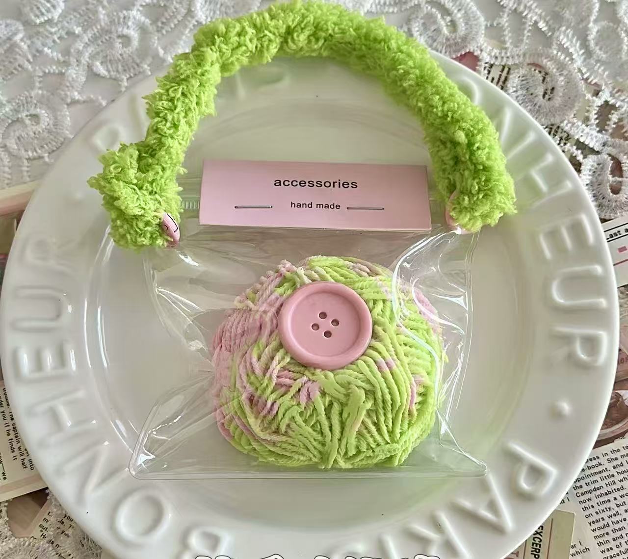 A 130g yarn ball squishy in pastel green and pink with a large pink button in the center, packaged in a transparent pouch with a fuzzy green handle.