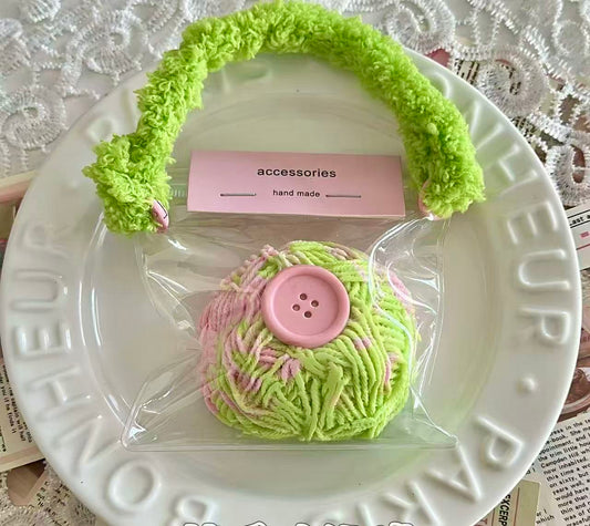 A 130g yarn ball squishy in pastel green and pink with a large pink button in the center, packaged in a transparent pouch with a fuzzy green handle.