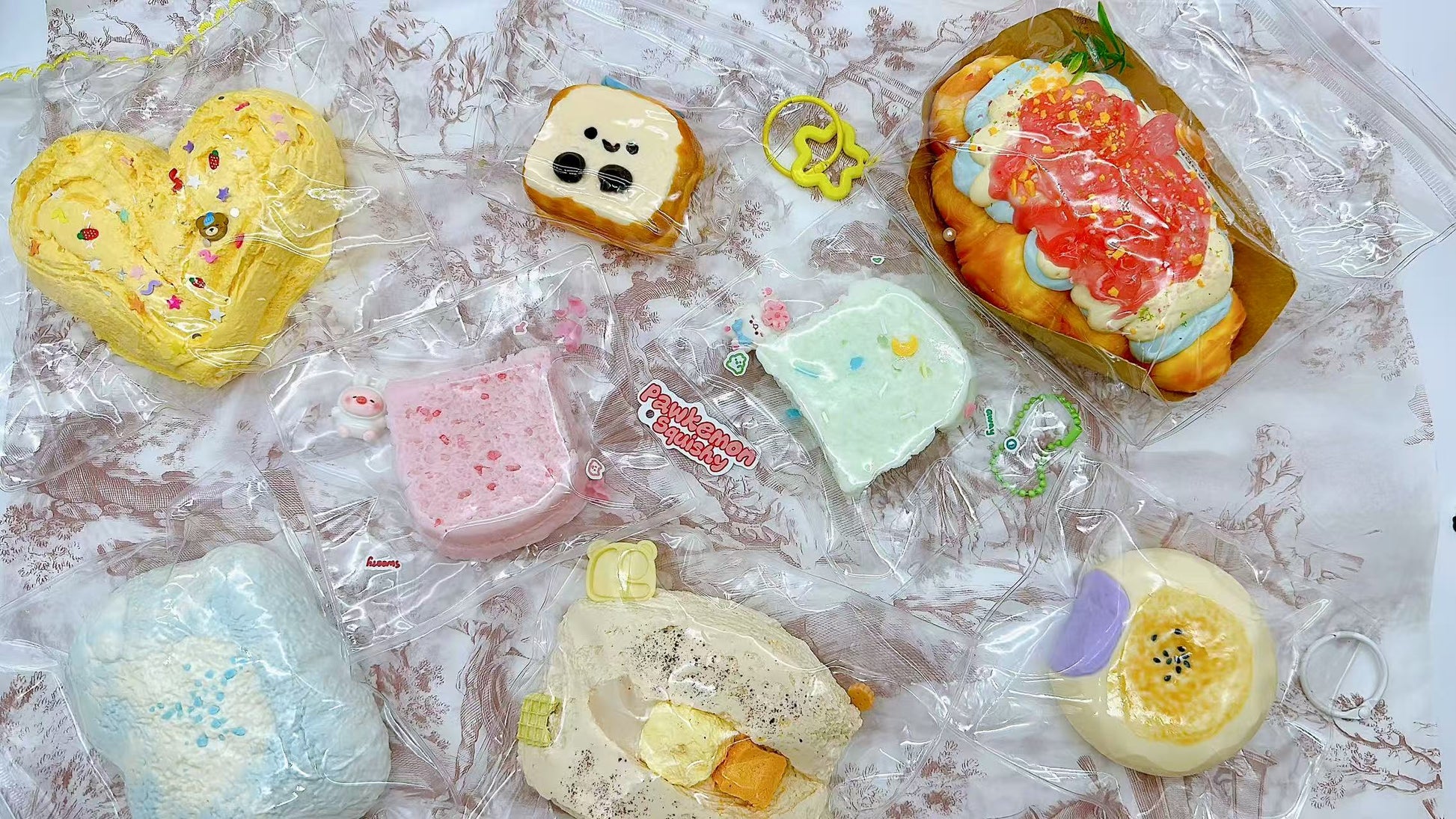  Variety of Pawkemon blind bag squishies laid out, including toast, decorated cream buns, pastel-colored bread slices, and textured clouds, all individually packaged with cute charms.