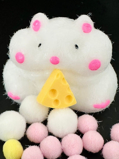 Close-up front view of cheese stealing hamster squishy toy with pastel pink cheeks, holding a cheese slice, surrounded by colorful balls