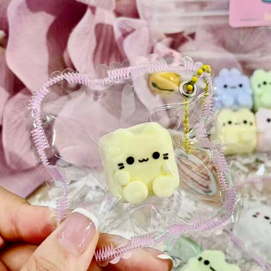 Handheld Custard Cube Cat Taba Squishy with tiny rounded limbs, soft custard color, and frilled packaging, perfect as a cute bag charm.
