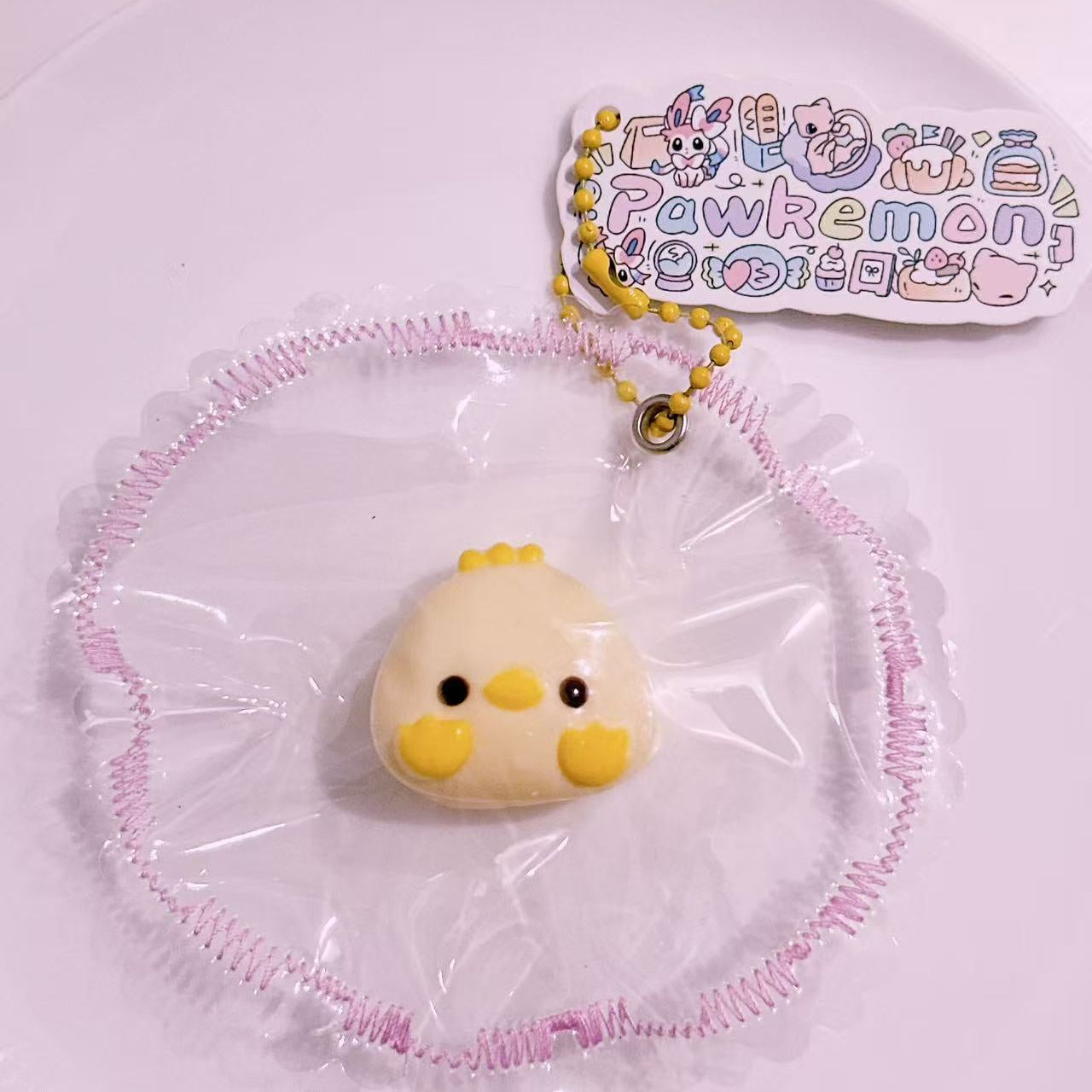 Eggie Chick Taba Squishy in protective packaging with yellow cheeks and soft pastel details. Cute handmade silicone squishy by Pawkemon.
