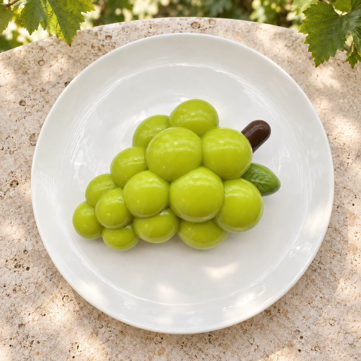 Alternate angled view of a handmade green grape Taba squishy resting on a white ceramic plate in soft outdoor sunlight.