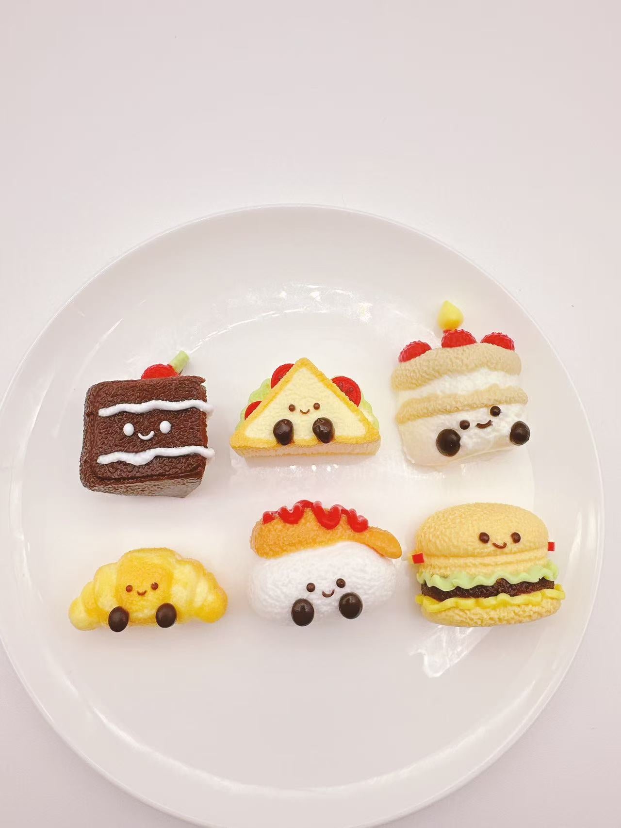 Six jellycat inspired food squishies displayed on a white plate, including cake, sandwich, croissant, shrimp tempura, burger and birthday cake.
