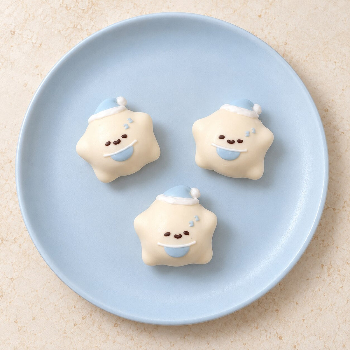 Three cream mini star Taba Squishies with blue nightcaps, crescent moons and cute faces on a light blue plate.