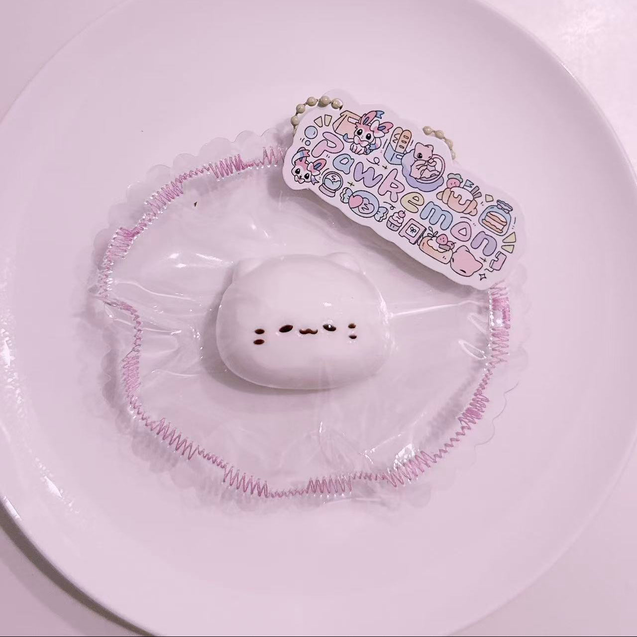 Mochi Meow Taba Squishy displayed on a plate with pastel frilled packaging and Pawkemon branding tag, featuring a soft mochi cat design.
