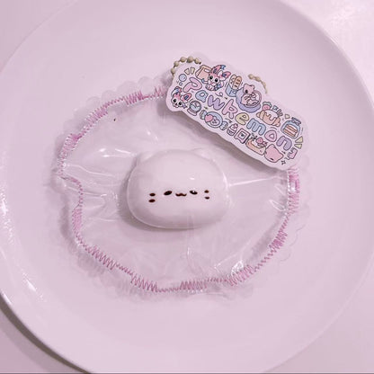 Mochi Meow Taba Squishy displayed on a plate with pastel frilled packaging and Pawkemon branding tag, featuring a soft mochi cat design.
