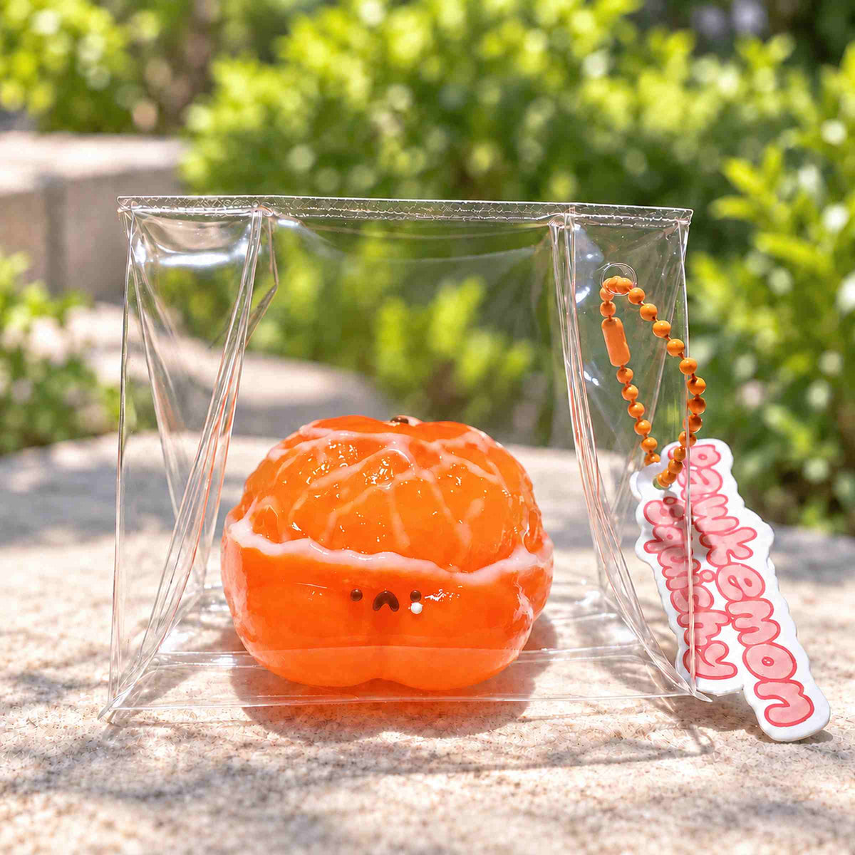 Orange hamster Taba squishy with glossy peeled citrus segments and a tiny face, displayed in a clear pouch on a sunlit stone surface. A Pawkemon Squishy tag hangs beside it.