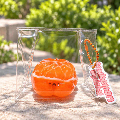 Orange hamster Taba squishy with glossy peeled citrus segments and a tiny face, displayed in a clear pouch on a sunlit stone surface. A Pawkemon Squishy tag hangs beside it.