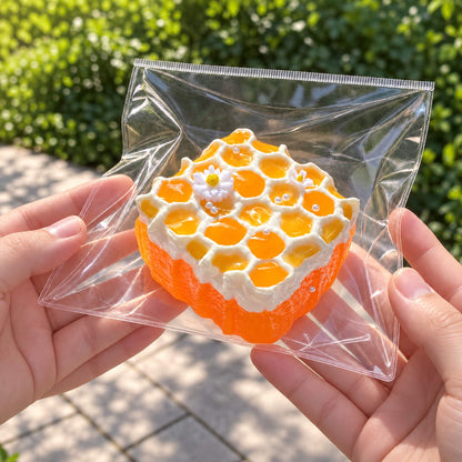 Two hands hold the honeycomb cake Taba Squishy in a clear bag against an outdoor garden background.