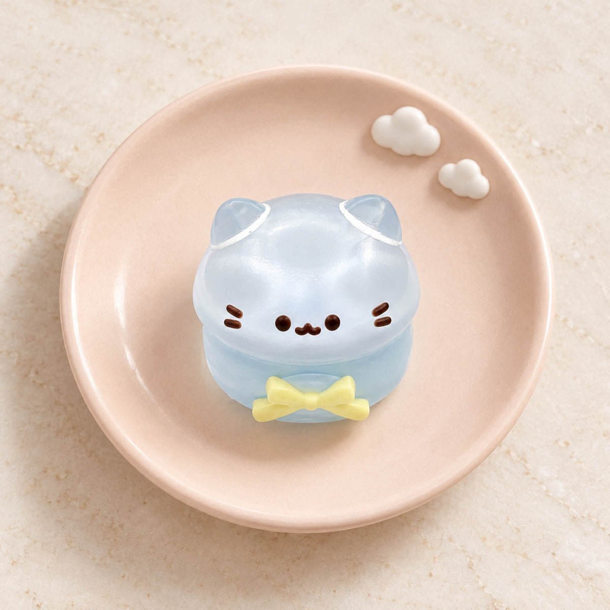 Clear pale-blue mini cat Taba Squishy with rounded ears, black whiskers and a yellow bow on a blush pink plate.